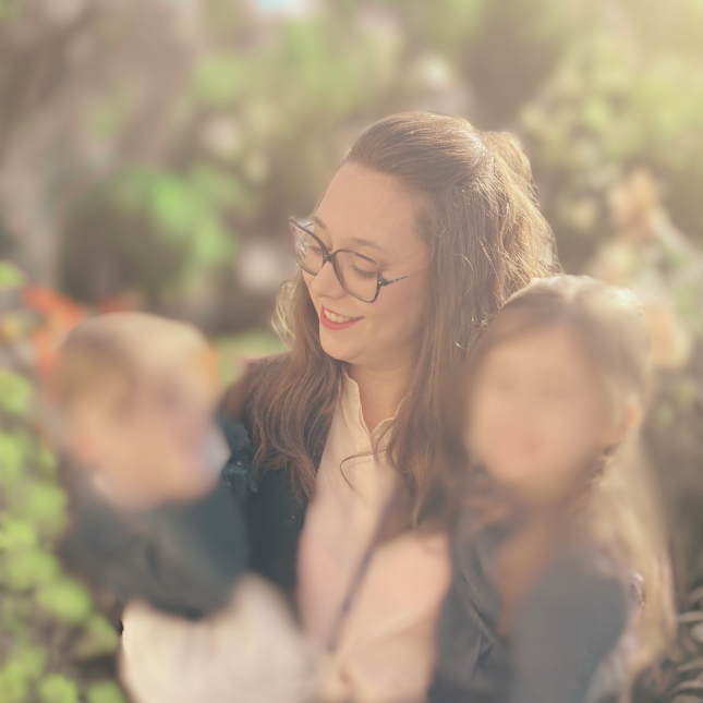 Maman souriante tenant ses deux enfants, entourée de fleurs floutées, et rayon de soleil en arrière-plan.